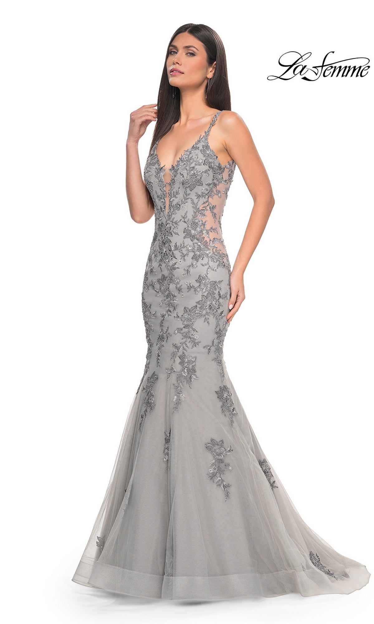 A woman wears the La Femme Backless Long Mermaid Prom Dress 32295—fitted, sleeveless, in silver-grey with floral lace and a mermaid silhouette—posing against a white background. The